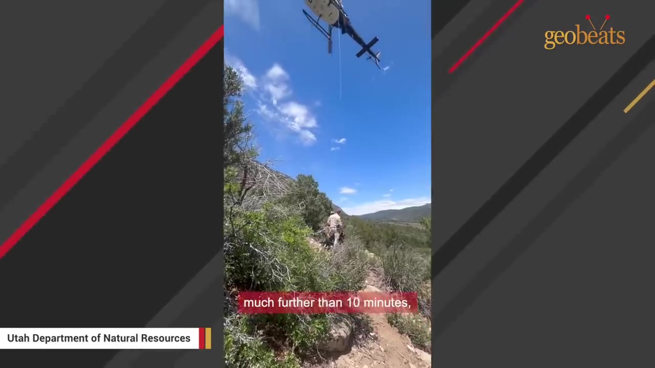A helicopter hovers in a bright blue sky above a wooded mountain trail where a rescue worker is walking.