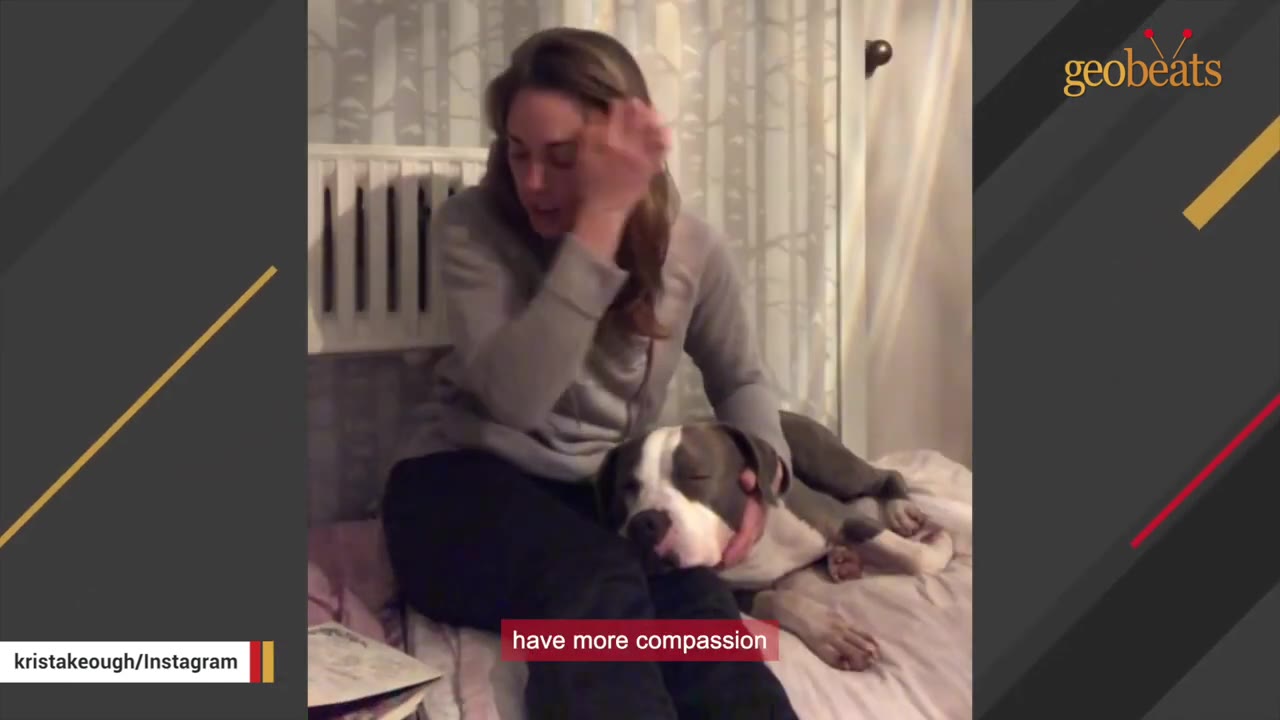 A woman sits on a bed, gently holding a grey and white dog that is resting its head on her lap.