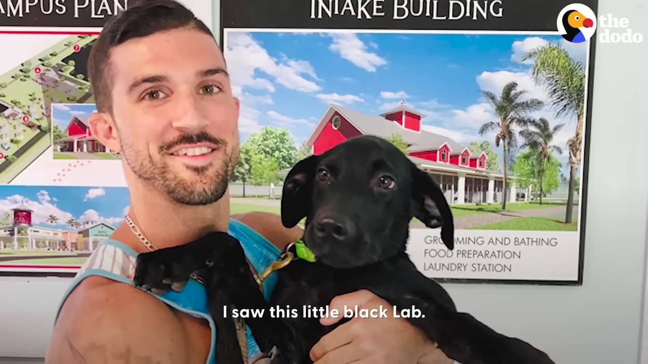 A smiling man holding a black Labrador puppy in front of an intake building sign.