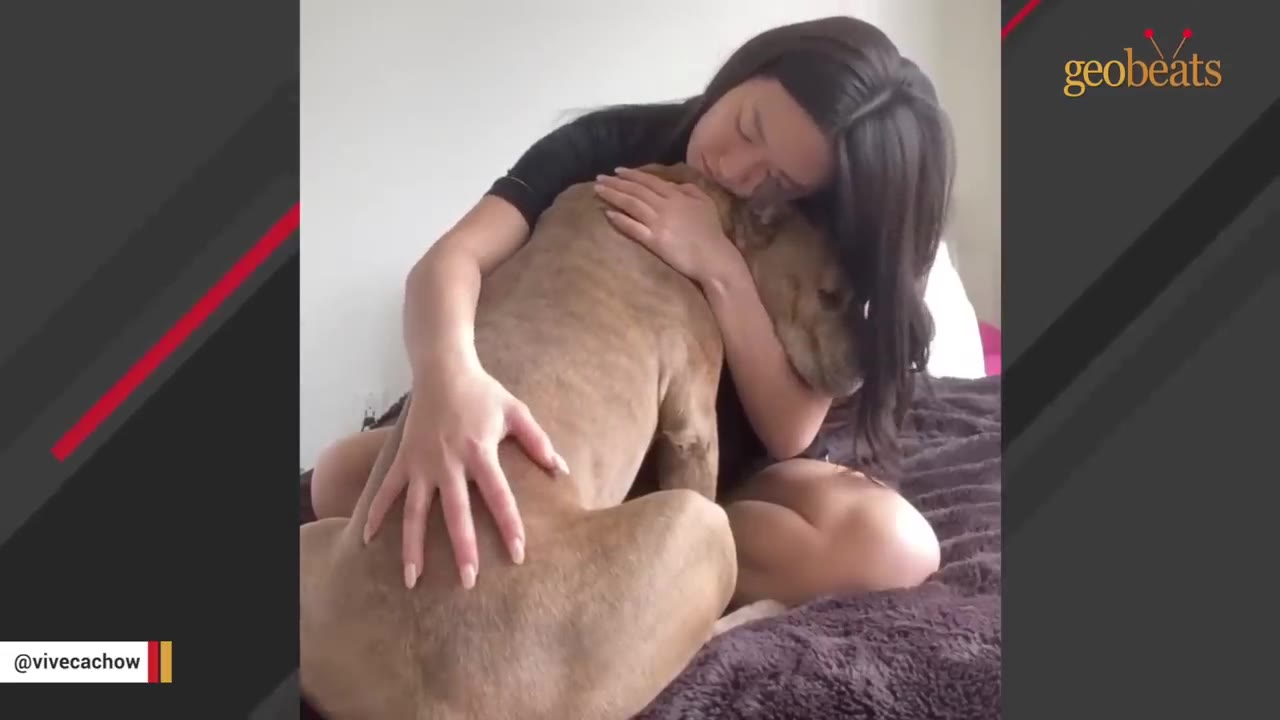 A woman sits on a bed, tenderly hugging a brown dog close to her chest.