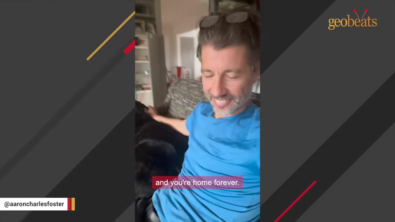 A smiling man in a blue t-shirt sits on a couch and pets a black dog next to him, with a subtitle that reads 'and you're home forever.'