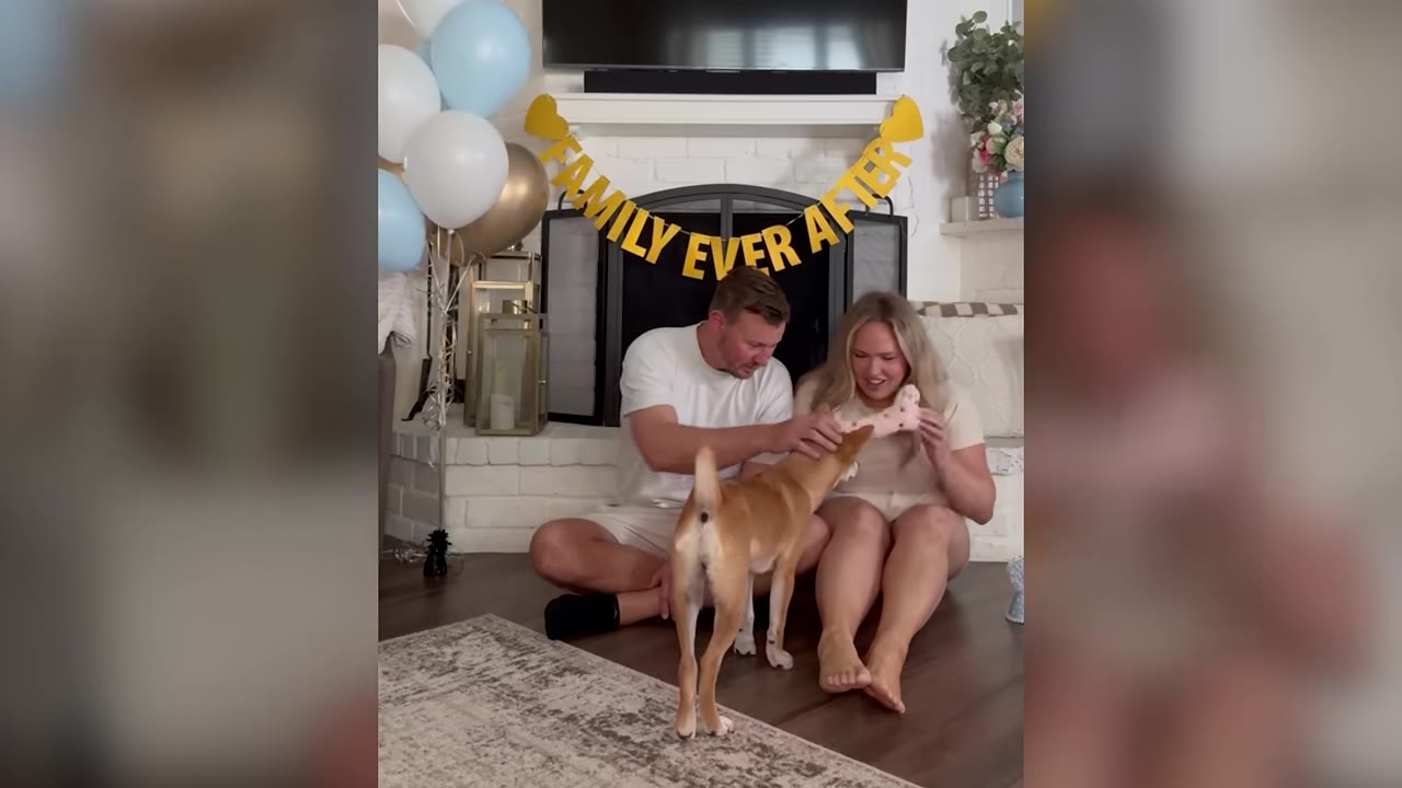 A man and a woman sit on the floor in front of a fireplace decorated with balloons and a banner that reads 'FAMILY EVER AFTER', smiling as they pet a tan dog standing in front of them.
