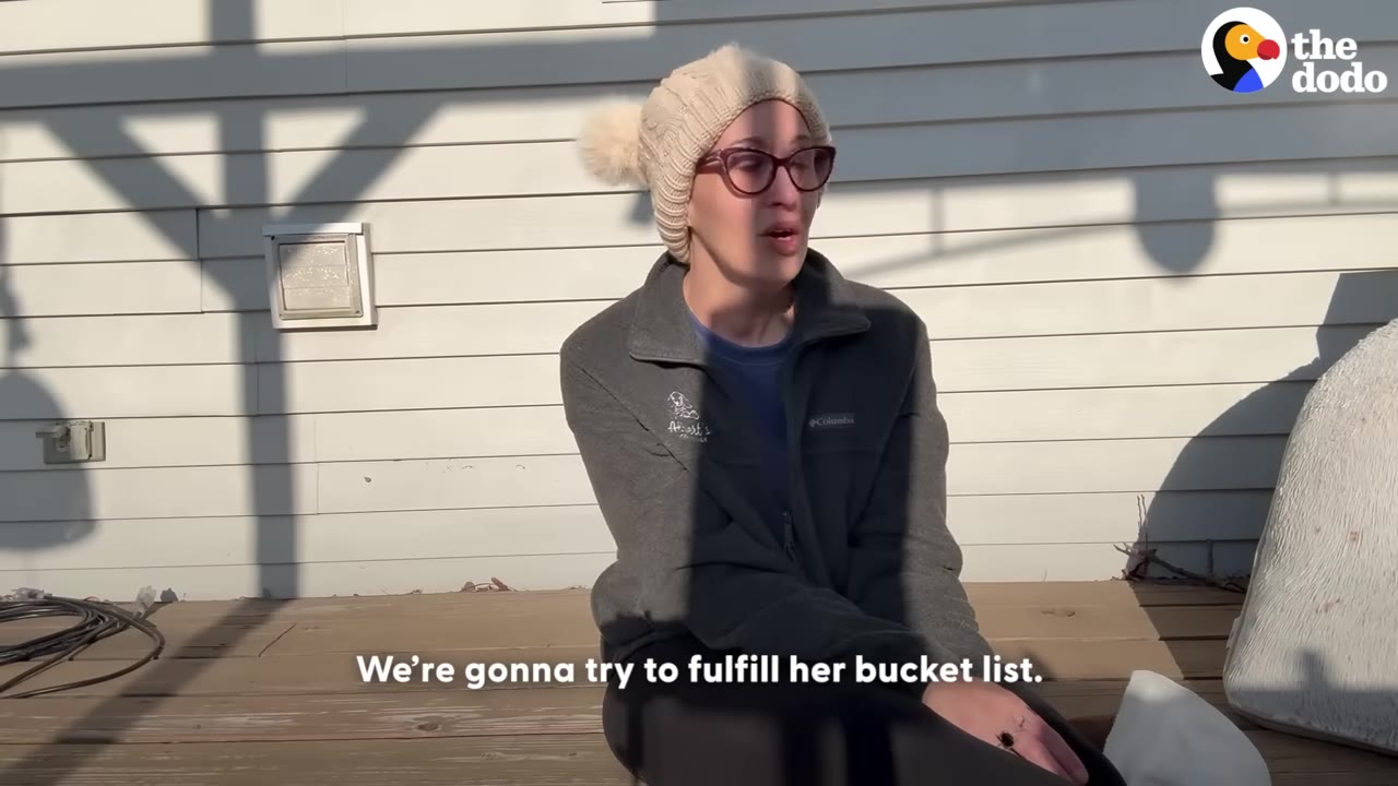 A woman wearing a knit hat and glasses sits outside on a wooden deck, speaking with an emotional expression.