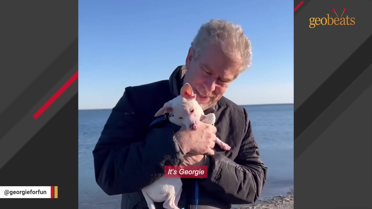 A man smiling warmly as he cradles a small white dog in his arms near a body of water.