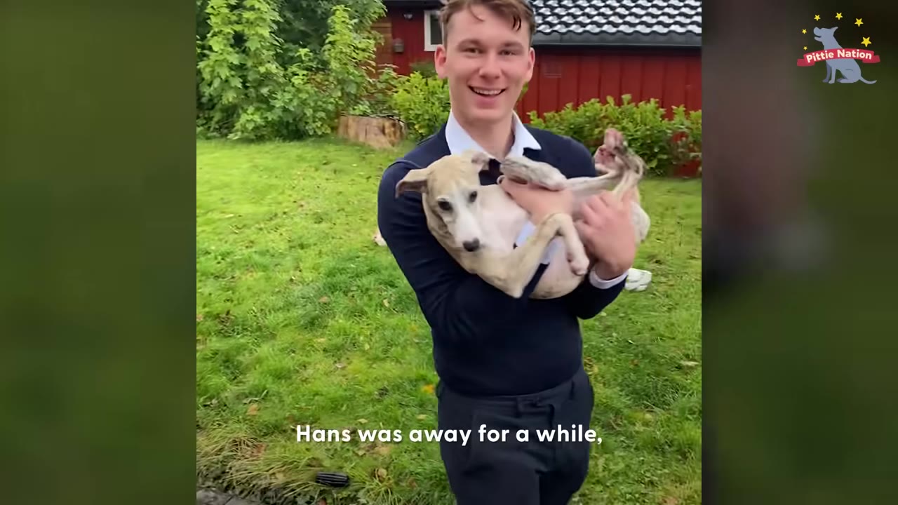 A smiling young man in a dark sweater holds a light-colored puppy in his arms while standing on a green lawn.