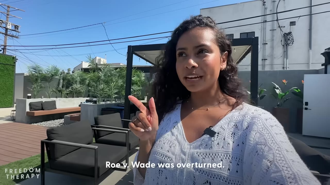 A woman with long curly hair stands on an outdoor patio, pointing her finger as she speaks, with the subtitle 'Roe v. Wade was overturned' displayed on the screen.