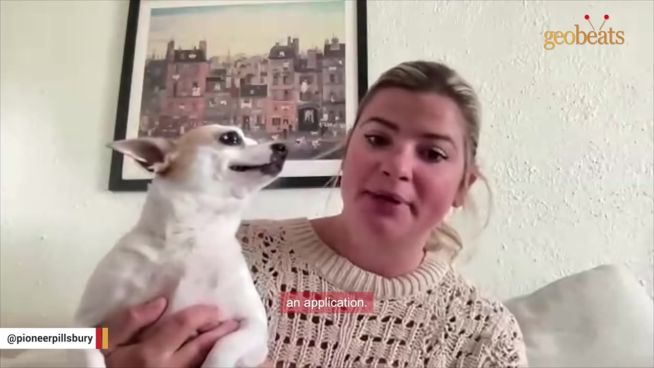 A woman with blonde hair tied back, wearing a cream-colored knit sweater, holds a small white and tan dog in her arms while speaking.