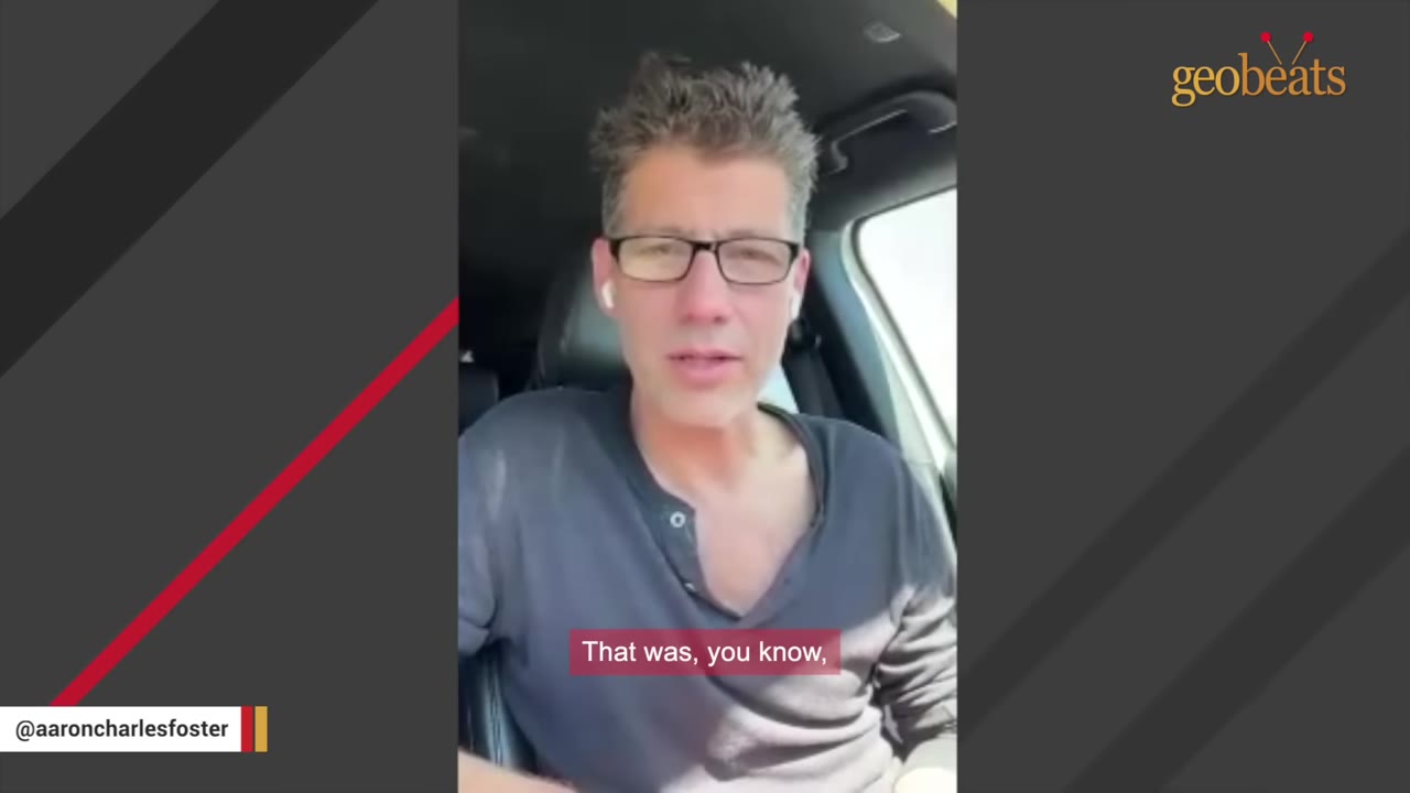 A man with glasses and a grey shirt speaks directly to the camera while sitting inside a car.