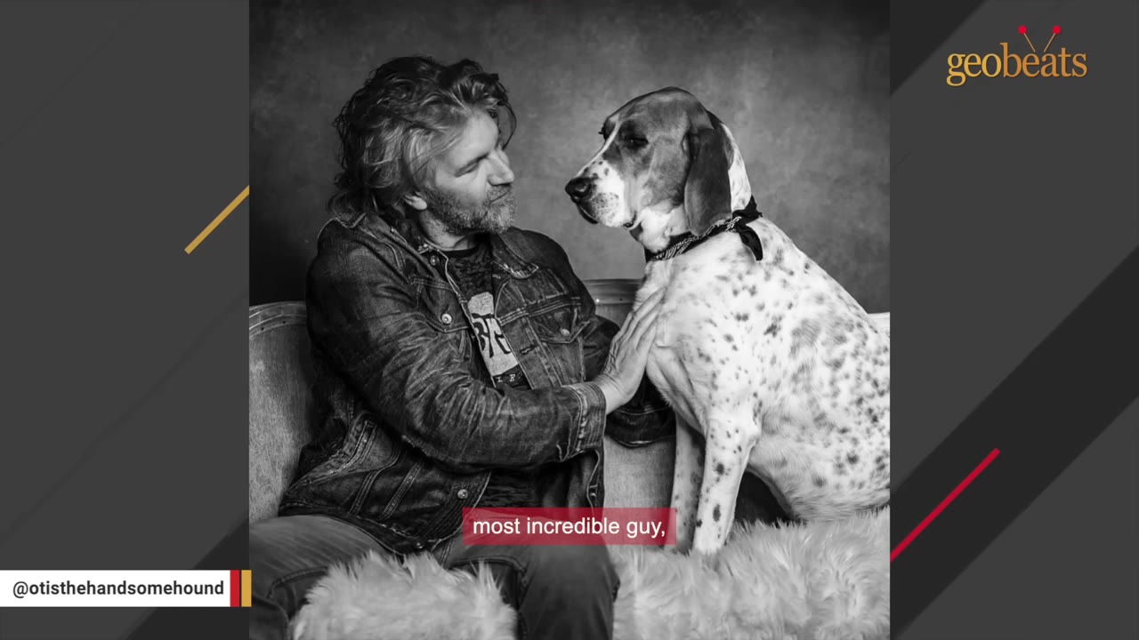 A black and white photo of a man with long hair and a beard, wearing a denim jacket, sitting on a couch and looking affectionately at a spotted hound dog.