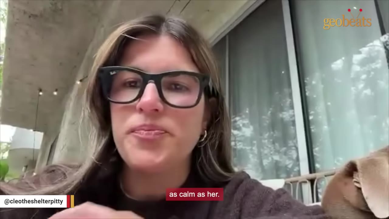 A close-up of a woman with glasses speaking to the camera, with a dog's head partially visible on the right.