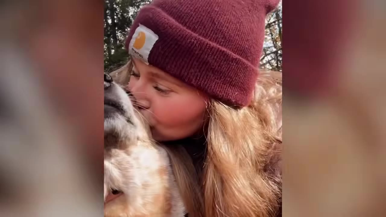 A close-up shot of a woman wearing a maroon beanie kissing the head of a light-colored dog.