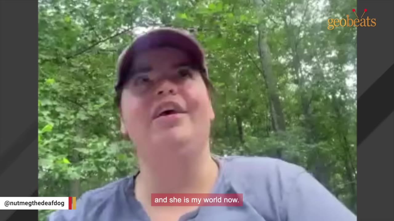 A close-up shot of a woman wearing a cap and a grey shirt, speaking outdoors with trees in the background.
