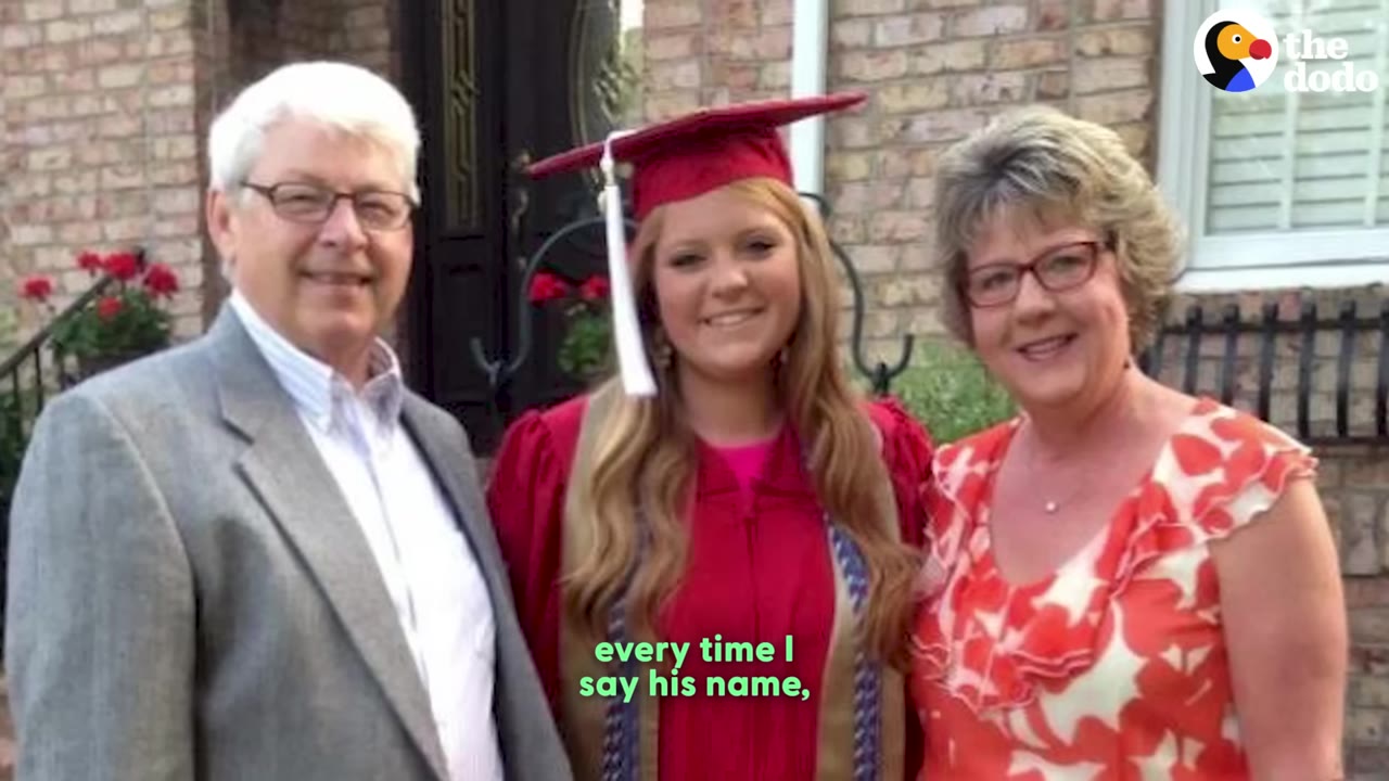 A family photo showing a man in a grey suit jacket, a young woman in a red graduation gown and cap, and a woman in a red and white floral dress smiling together in front of a brick house.