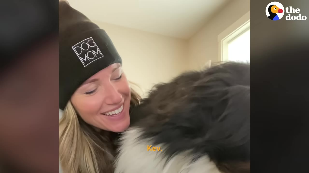 A smiling woman wearing a black beanie with "DOG MOM" printed on it is cuddled by a fluffy Bernese Mountain Dog.