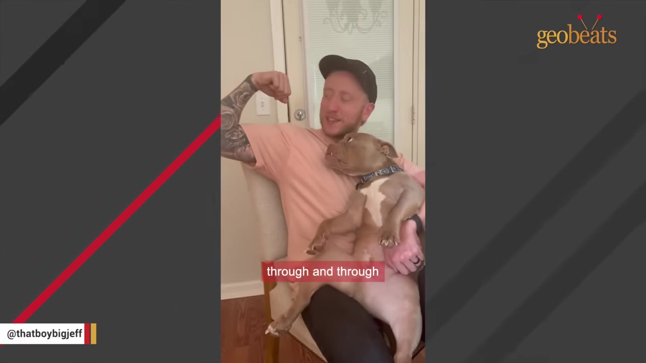 A man in a pink shirt and black cap sits in a chair, holding a light brown dog on his lap.