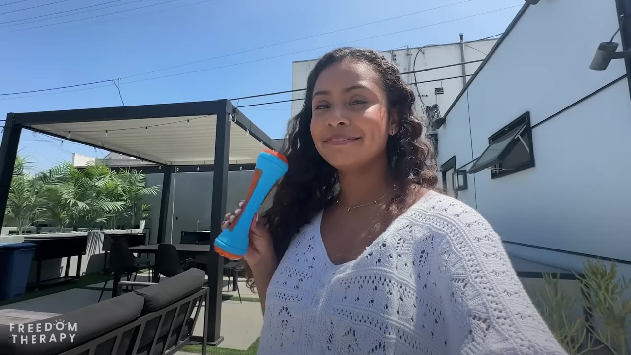 A smiling woman with long curly dark hair holds up a blue and orange dog toy in an outdoor patio setting.
