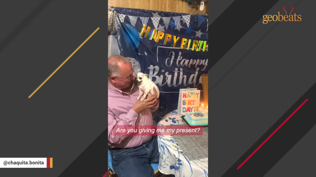 A man in a pink checkered shirt holding a small white Chihuahua close to his face, looking at it affectionately in front of a birthday banner.