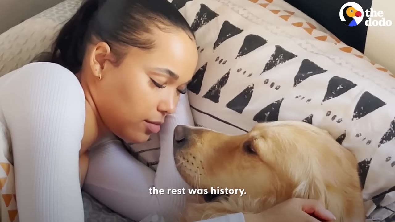 A woman with her hair tied back lies in bed, facing a golden retriever dog resting its head near hers on a patterned pillow.