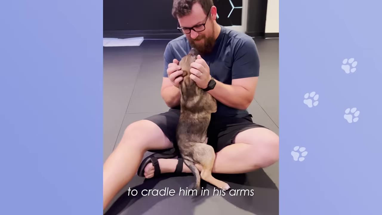 A man with glasses and a beard sits cross-legged on a dark floor, cradling a small brown puppy in his arms as the puppy nuzzles his face.