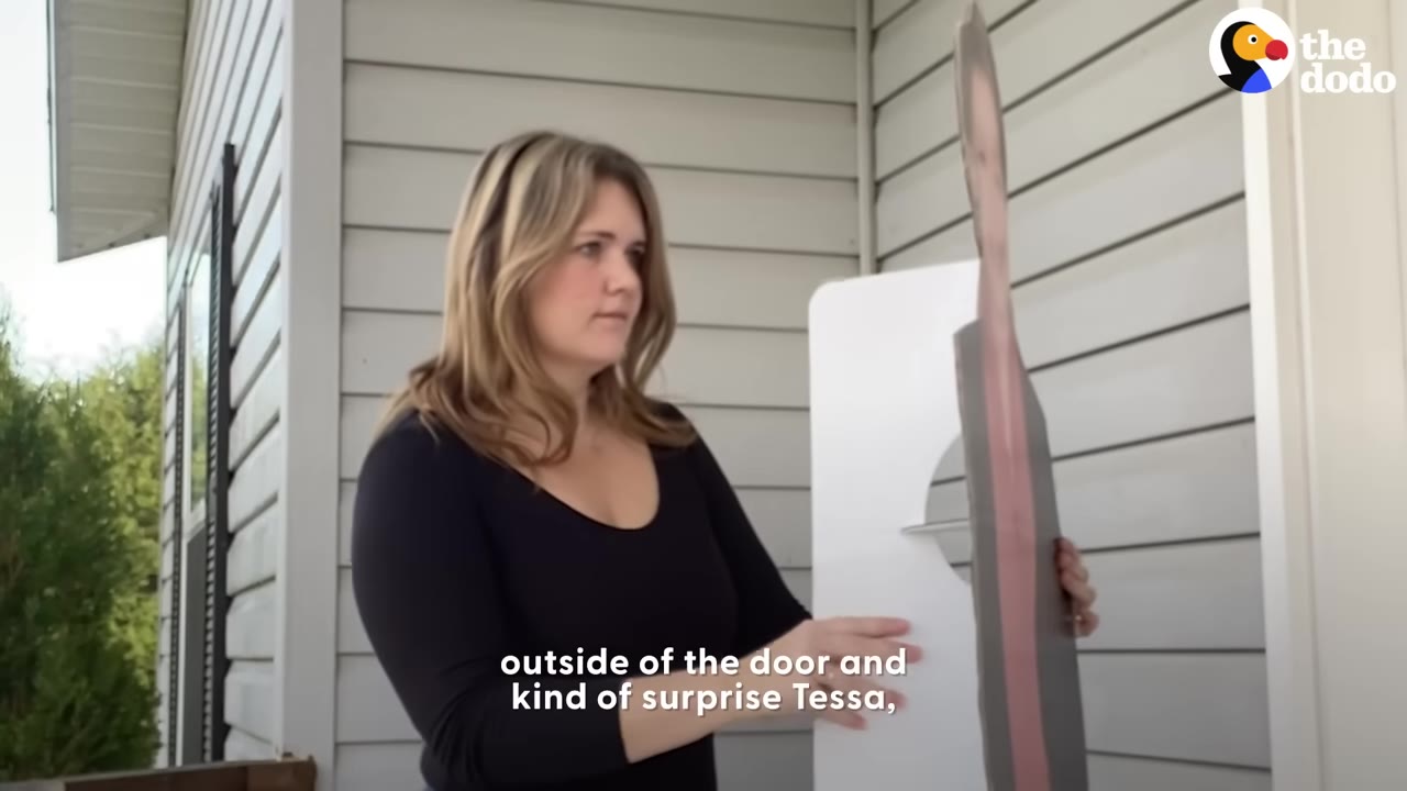 A woman stands outside a house holding a life-size cardboard cutout of a person.
