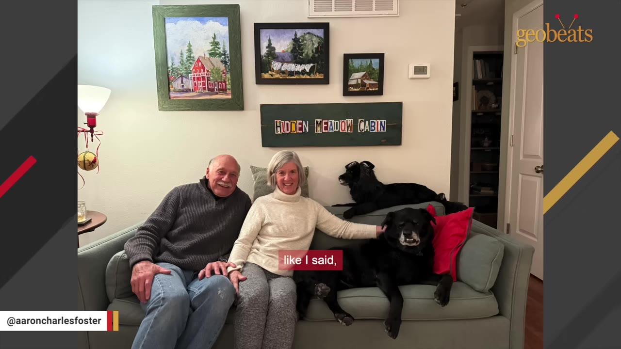 An older man and woman sit smiling on a light green couch alongside two black dogs, with framed paintings hanging on the wall behind them.