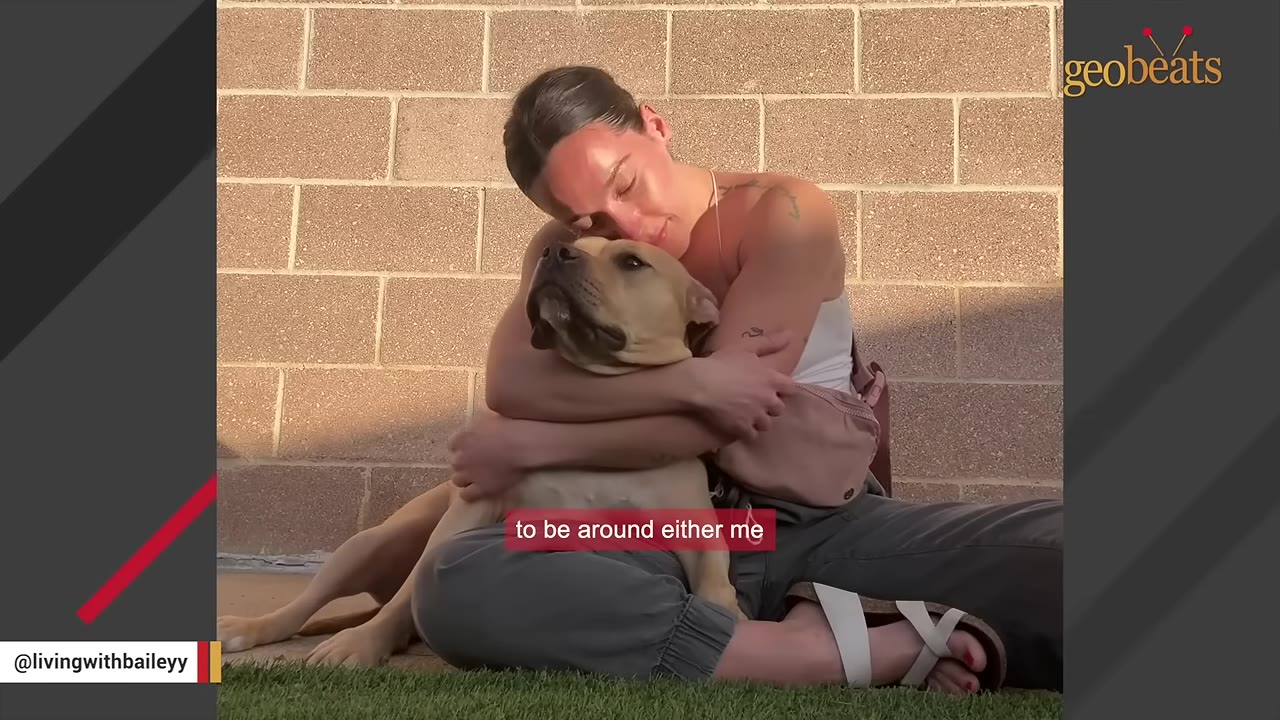 A woman sitting cross-legged on the grass hugs a light brown dog tightly against her chest.