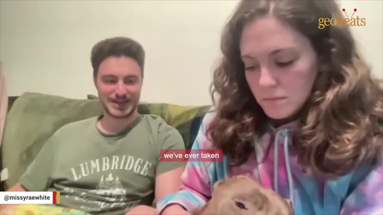 A man and a woman sitting on a couch, looking down affectionately at a dog they are holding.