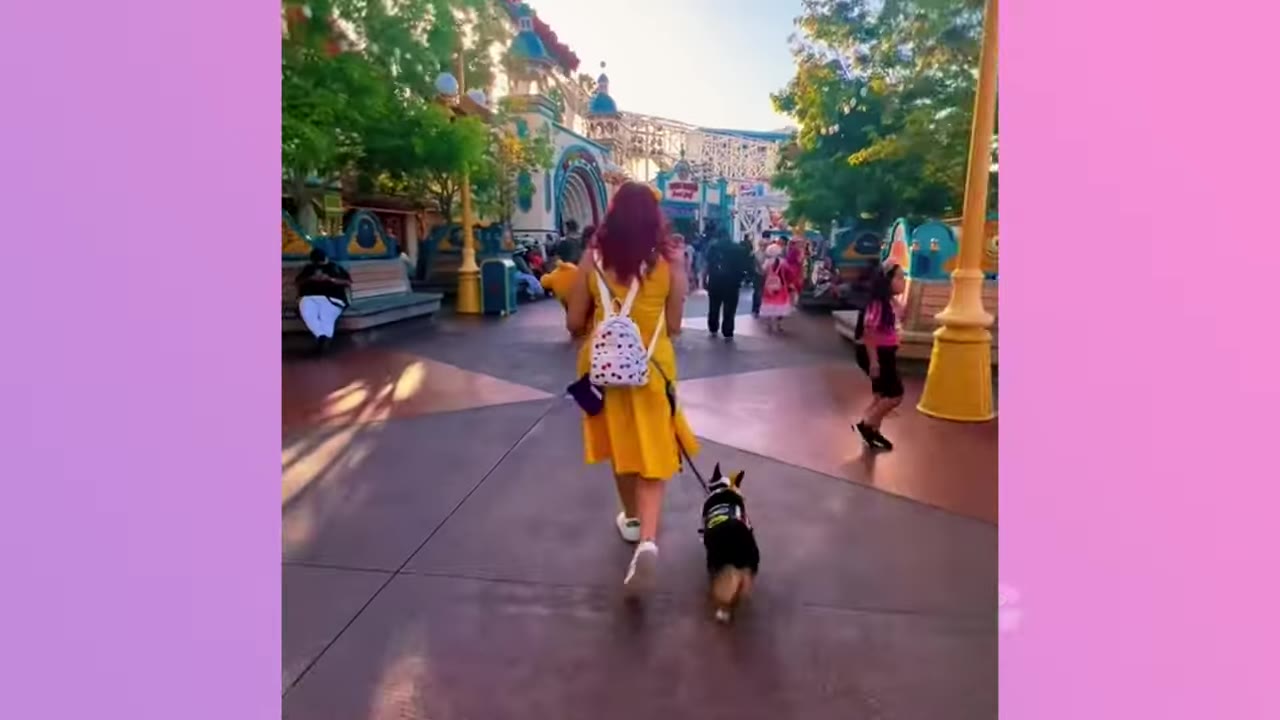 A woman with red hair wearing a yellow dress walks her corgi on a leash through a theme park plaza with other visitors in the background.