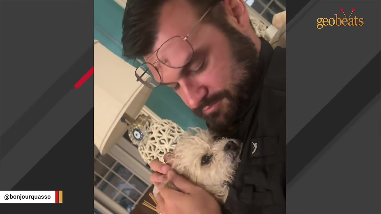 A man with glasses and a beard cradles a small, light-colored scruffy dog close to his chest.