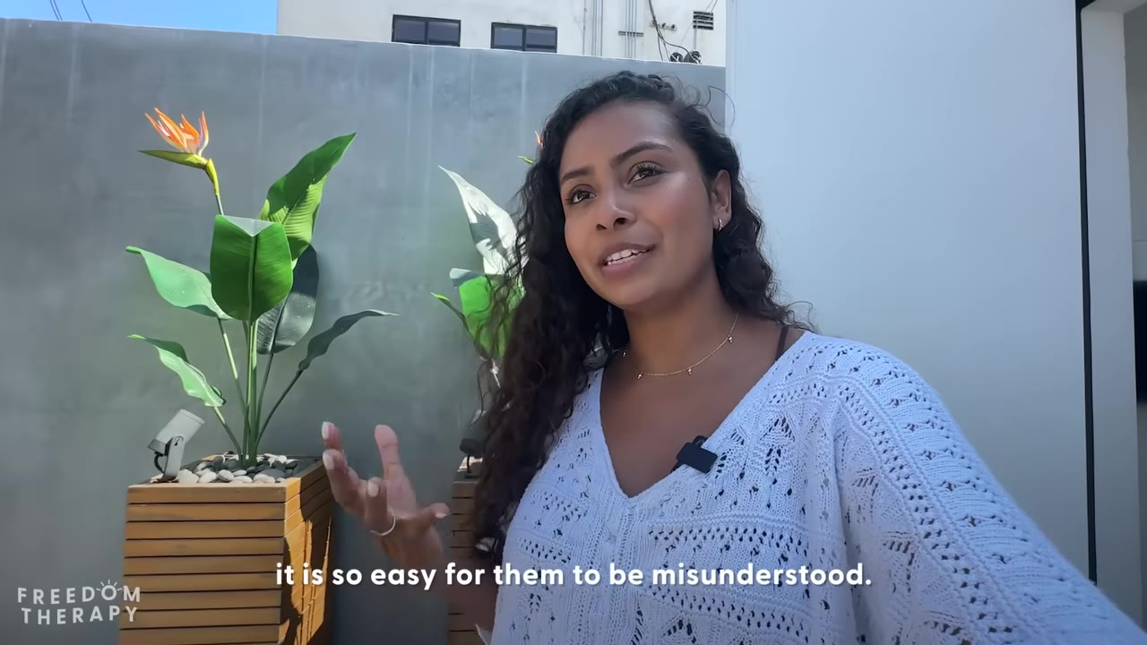 A woman with curly hair wearing a white knit top speaks outdoors, gesturing with her hand, with a subtitle that reads 'it is so easy for them to be misunderstood.'