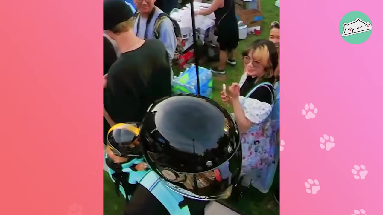 A smiling woman in a crowd holds up her phone to take a photo of a motorcyclist wearing a black helmet.