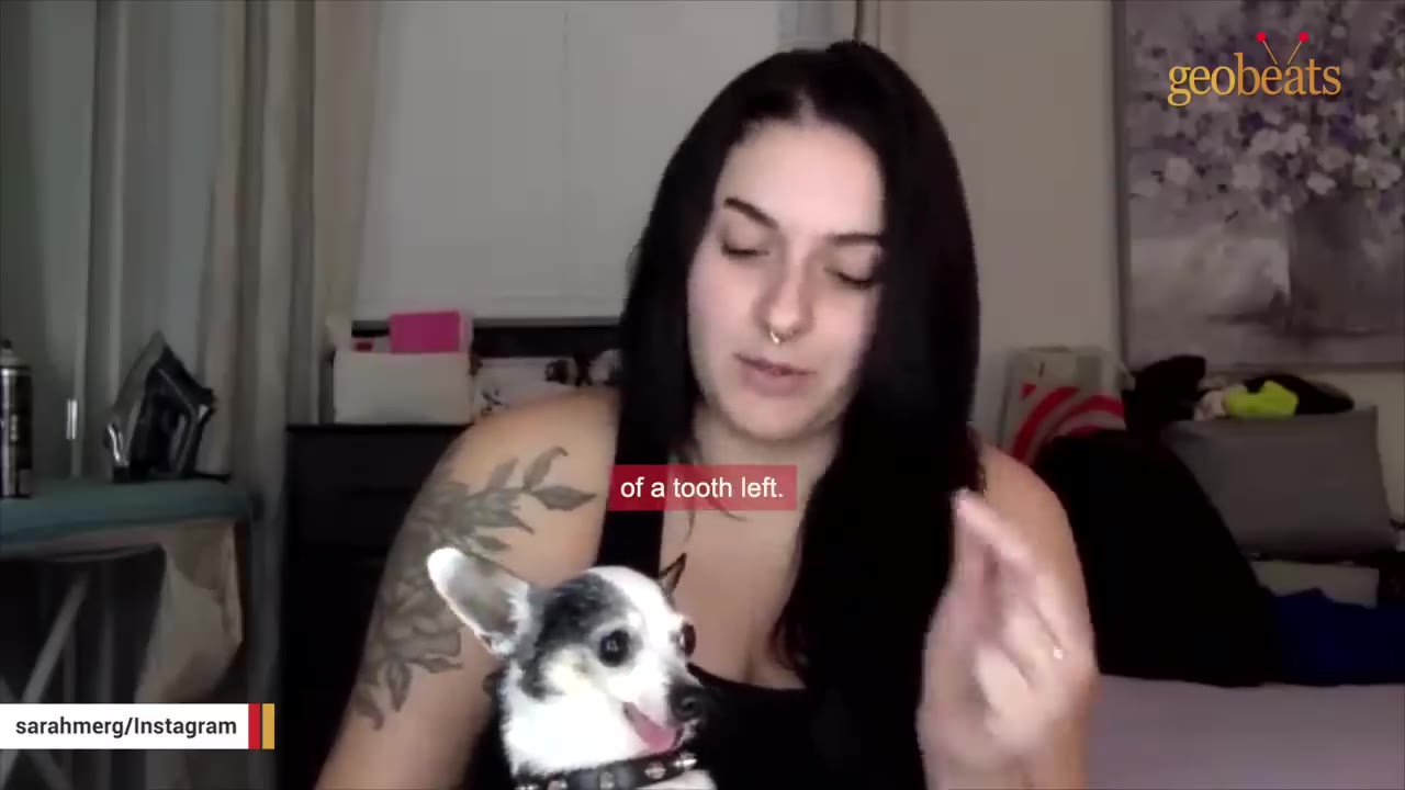 A woman with dark hair and a shoulder tattoo holds a small black and white chihuahua whose tongue is sticking out.
