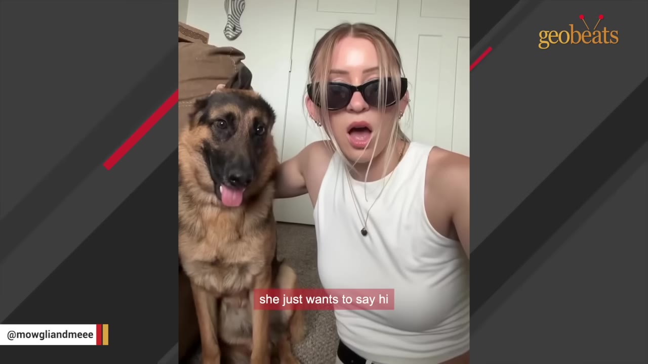 A German Shepherd sits next to a woman wearing sunglasses and a white tank top, who is smiling and gesturing towards the camera.