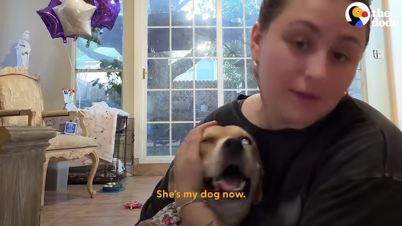 A woman holds a light brown dog close to her face as she speaks, with the subtitle 'She's my dog now' on the screen.