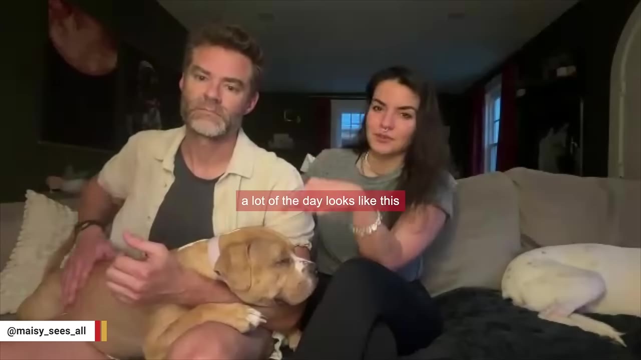 A man and a woman sit on a couch, with the man holding a light brown dog in his lap while petting it.