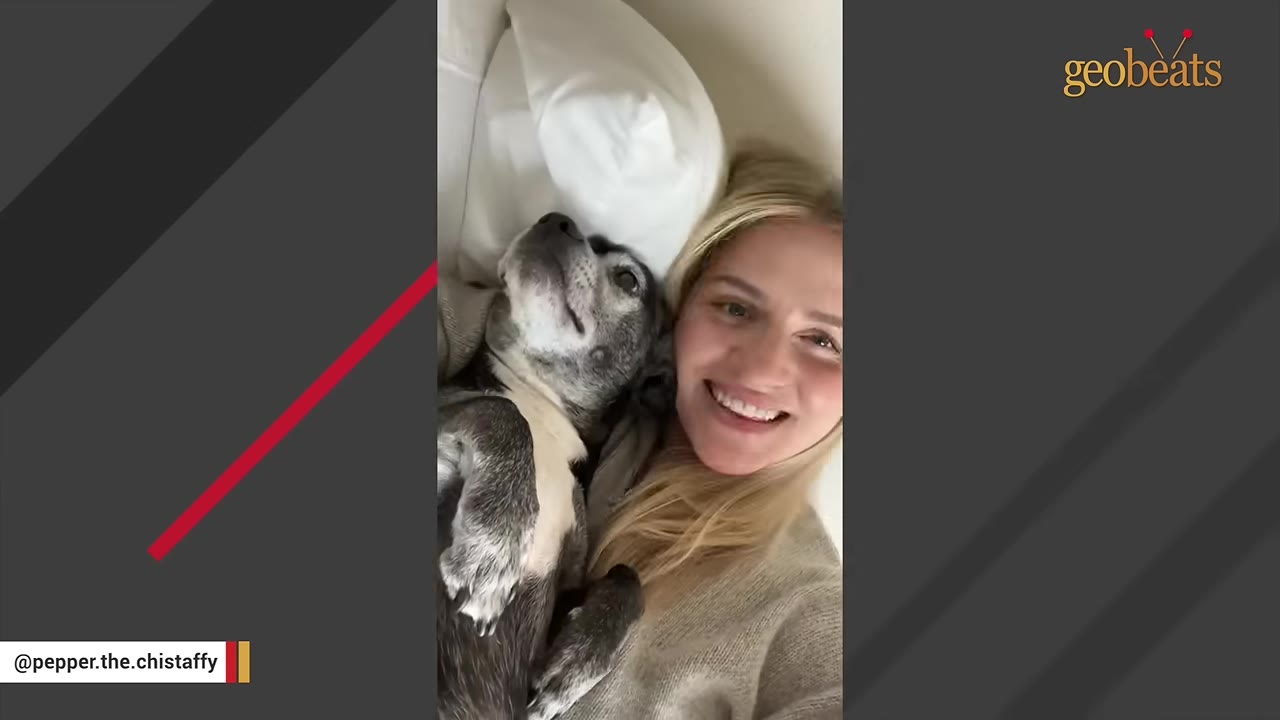 A smiling blonde woman lying down next to her grey and white dog resting its head on a pillow.
