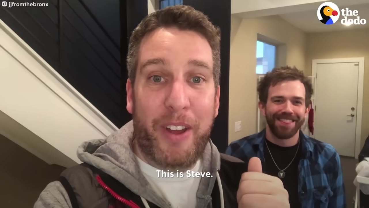 A man in a grey hoodie smiles at the camera, pointing his thumb back toward another smiling man in a blue plaid shirt.