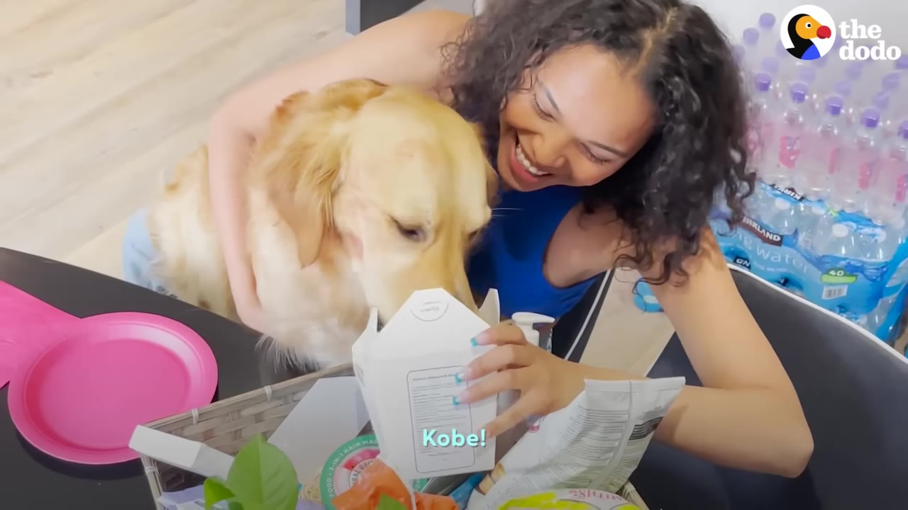 A smiling woman with curly hair hugs a golden retriever while holding open a white gift box filled with treats.