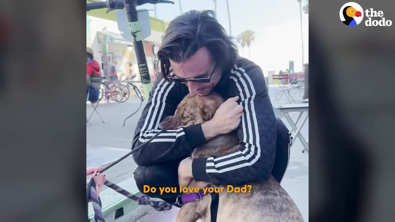 A man wearing a black track jacket with white stripes hugs a brown dog closely while kneeling outdoors.