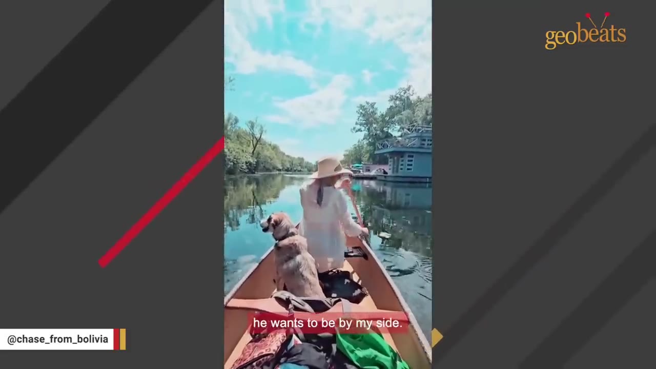 A dog sits in the front of a canoe facing a woman in a sun hat who is paddling down a calm river.