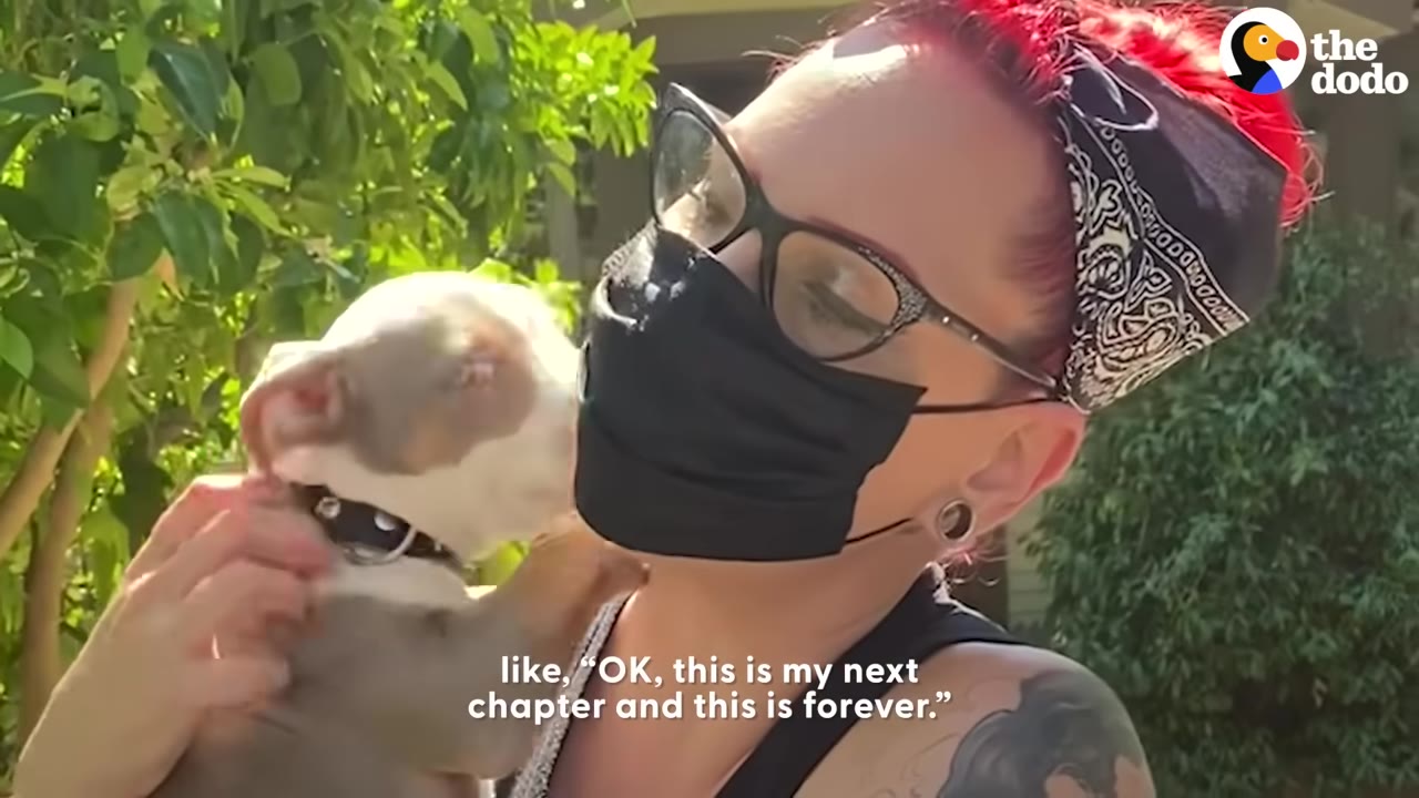 A woman with red hair, glasses, and a black face mask holds a small tan and white puppy close to her face as the puppy nuzzles her cheek.