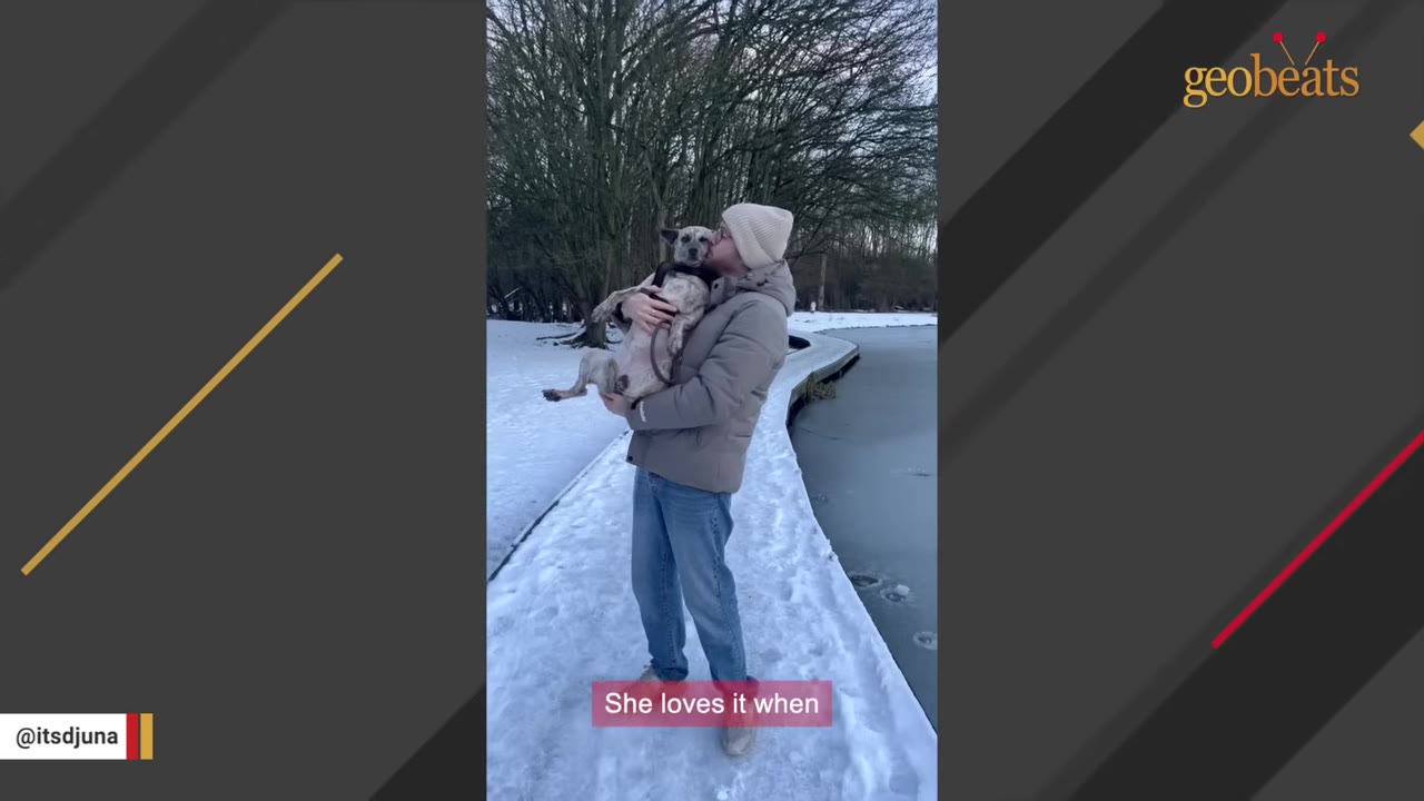 A man in a winter jacket and beanie holds a medium-sized grey dog in his arms and kisses her on the head while standing on a snow-covered path next to a frozen body of water.