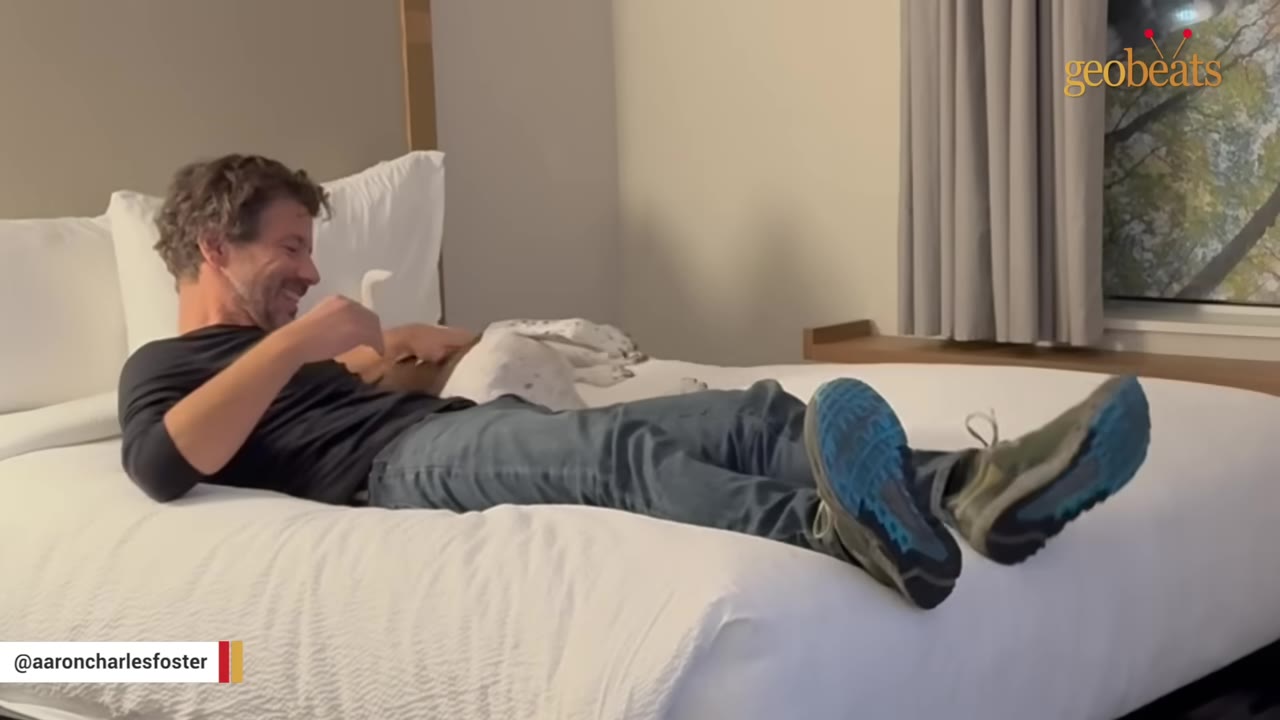 A man lying on a white bed, smiling at a white and black spotted dog resting beside him.