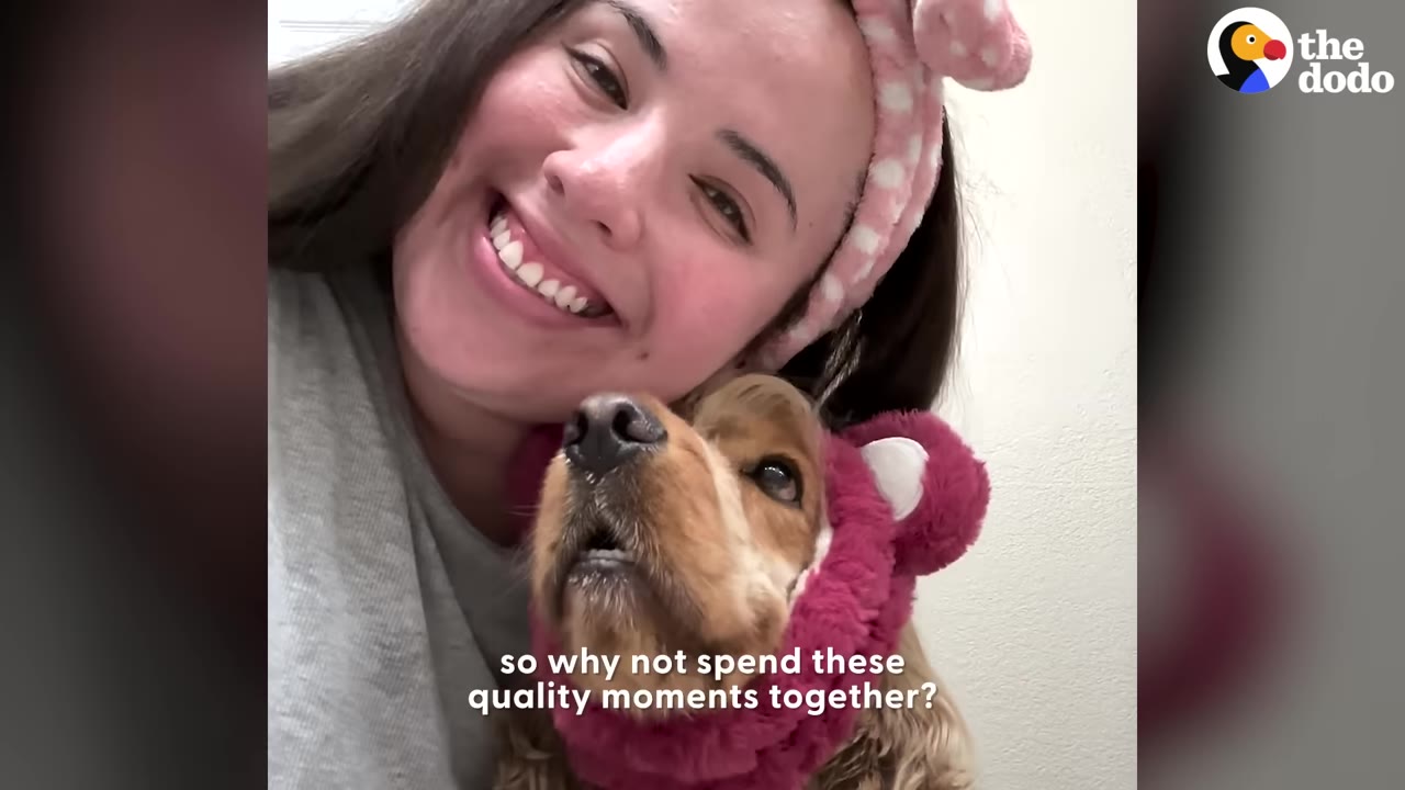 A smiling woman with a pink headband leans her head against her cocker spaniel dog, who is wearing a fuzzy pink bear-ear hood.