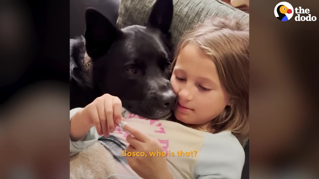 A black dog rests its head affectionately against a young girl's cheek as she holds him close.