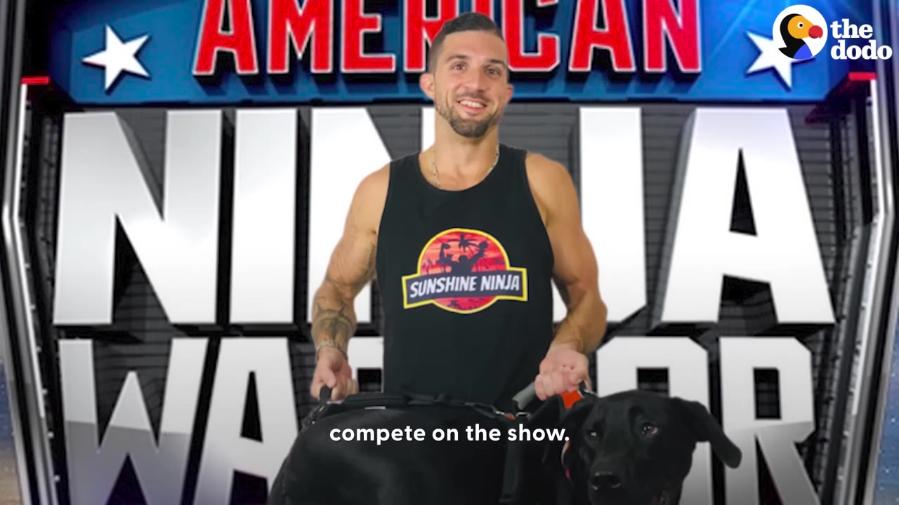 A smiling man in a black tank top stands in front of a large 'American Ninja Warrior' sign, holding his black dog.
