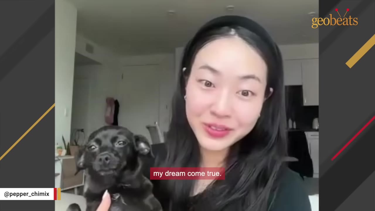 A smiling woman with long dark hair holds a small black dog in her arms as she speaks.
