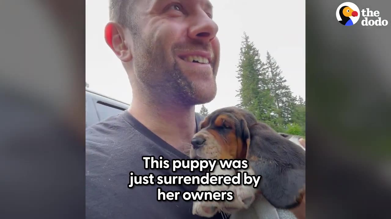 A man holding a small hound puppy with long, floppy ears close to his chest outdoors.