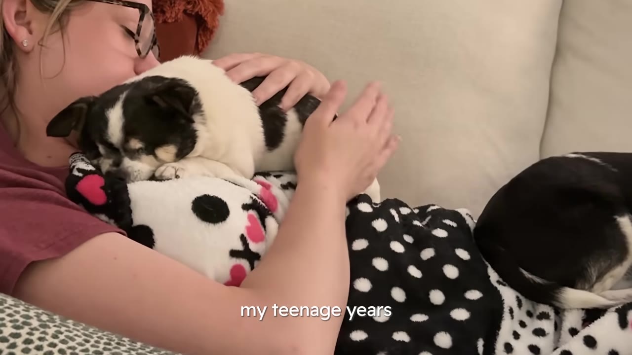 A woman lying on a couch gently hugging and cuddling her small black-and-white dog.