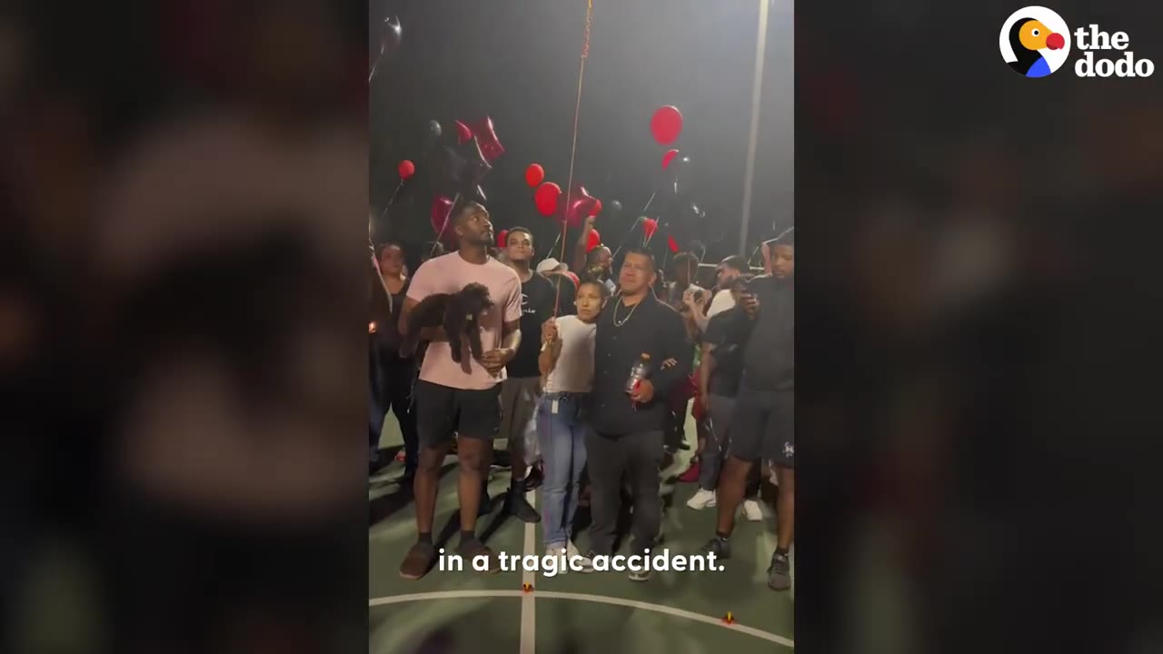 A group of people gathered at night on an outdoor court holding red balloons during a memorial gathering.